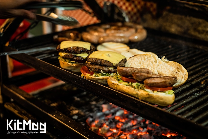 انواع یرگر های دوبل برگر که روی گریل ذغالی قرار گرفته همچنین پنیر ورقه ای پیتزا سوسیس ژامبون و نان های باگت و برگری قرار گرفته است