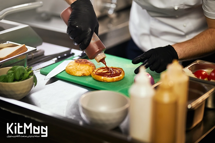 یک آشپز در حال سس زدن به نان برگر می باشد که روی میز رستوران قرار گرفته است