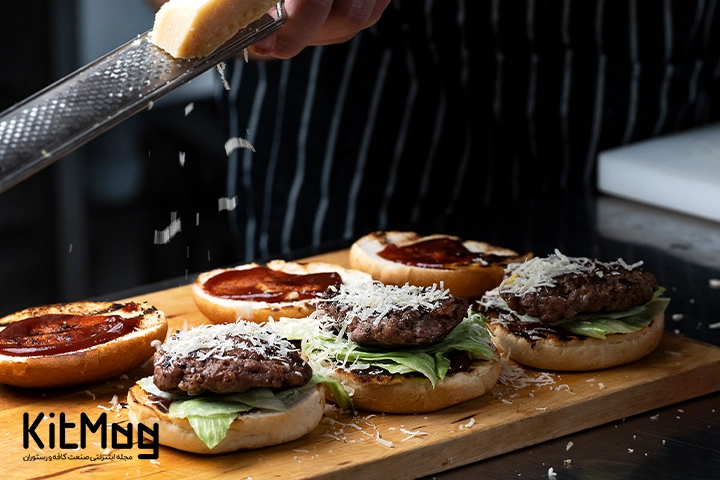 سه عدد همبرگر که آشپز در حال سس زدن روی آن ها می باشد و روی تخته چوبی قرار گرفته اند