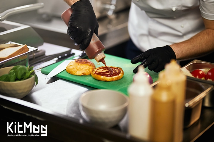 یک آشپز در حال ریختن سس قرمز روی لایه زیرین نان همبرگر می باشد با سس پمپی که روی یک تخته گوشت قرار گرفته است و در آشپزخانه رستوران است
