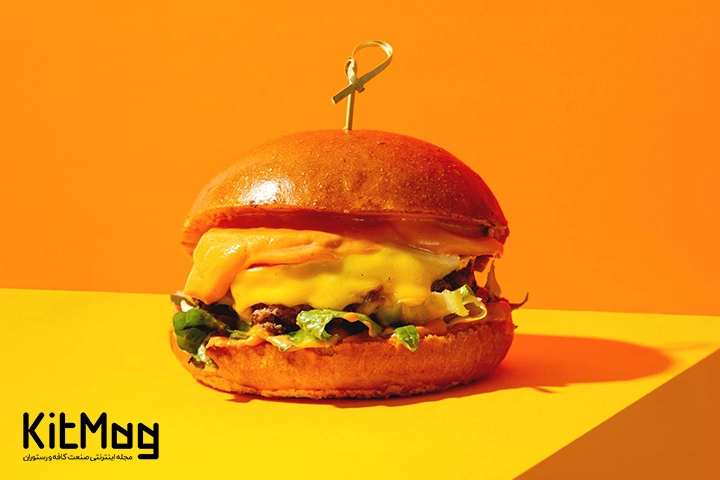 عکس یک چیزبرگر که رنگ پس زمینه آن نارنجی می باشد و برگر روی یک میز قرار گرفته که به رنگ زرد رنگ است و پنیر همبرگر از آن بیرون زده است