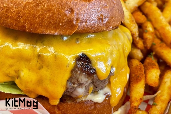 پنیر چداری که از لابه لای همبرگر چیزبرگر بیرون ریخته و سیب زمینی در کنار آن قرار گرفته است از نمای نزدیک
