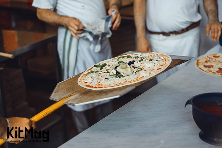 یک پیتزا ایتالیایی که روی یک پارو پیتزا قرار گرفته است و آشپز در حال قرار دادن آن در فر است