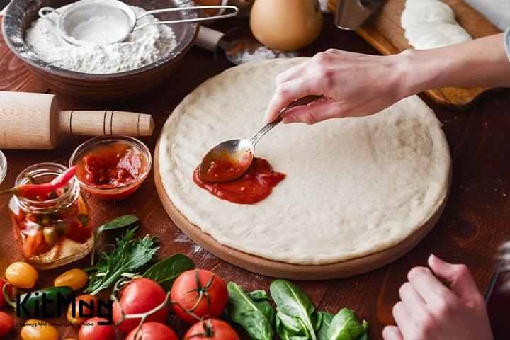 یک آشپز خانگی در حال پهن کردن سس پیتزا روی خمیر پیتزا می باشد