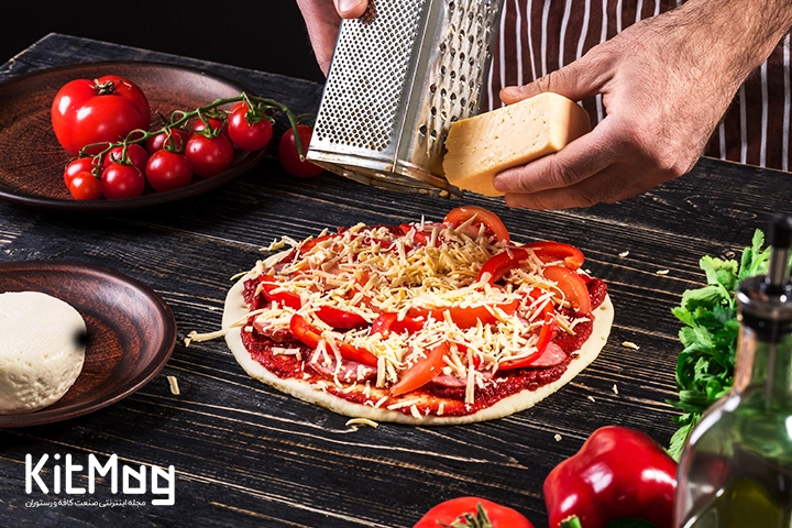 یک آشپز در حال رنده کردن پنیر پارمزان روی یک پیتزا با فلفل قرمز می باشد