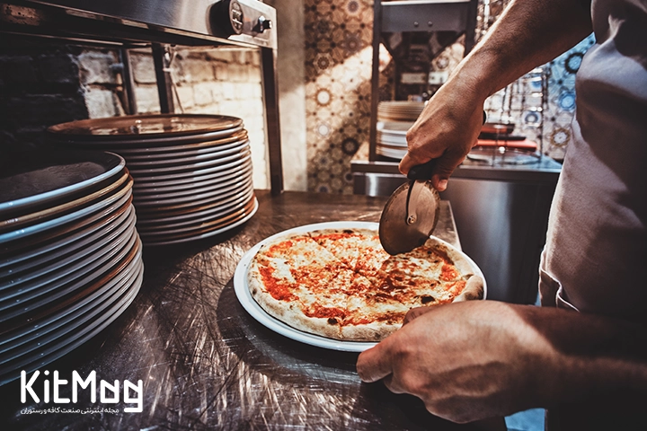 یک آشپز در حال برش پیتزا با کاتر رولی مخصوص می باشد که روی یک میز استیل رستوران قرار گرفته است