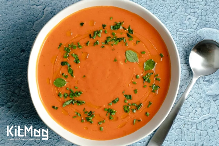 کاسهای از گاسپاچو که روی آن سبزیهای خردشده پاشیده شده است. کاسه روی سطحی آبیرنگ و خالدار قرار دارد و یک قاشق فلزی در سمت راست دیده میشود.