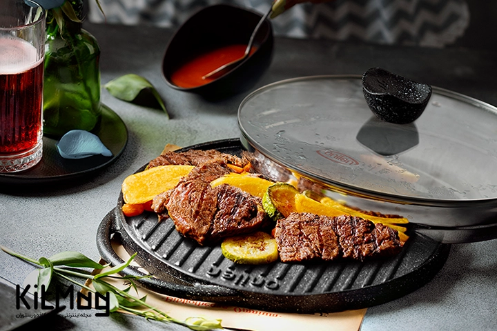 سینی یا تابه در دار برند نالینو Nalino که داخل آن گوشت استیک پخته شده است و کنار آن با نوشیدنی آبجو تزئین شده است و روی یک میز رستورانی شکیل به رنگ طوسی قرار گرفته است