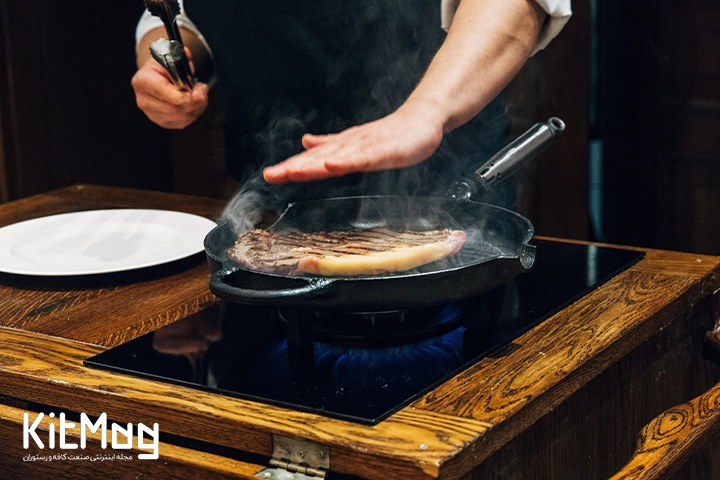 آشپز در حال پخت استیک داخل یک تابه چدنی می باشد و همچنین در فضای آشپزخانه مدرن یک رستوران مشغول آشپزی می باشد