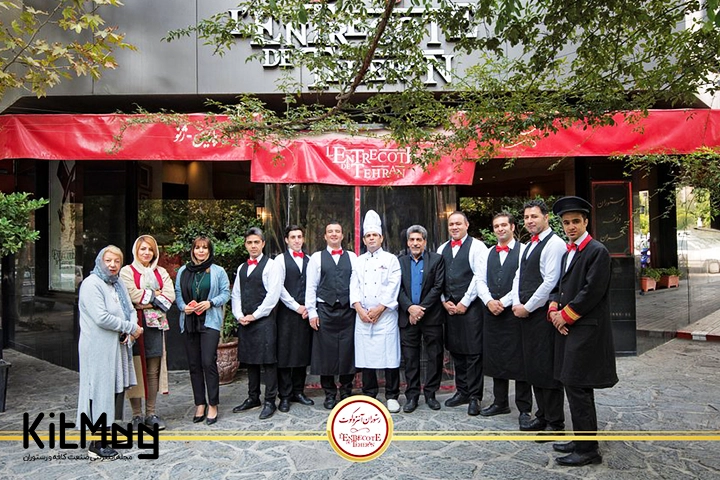 کارمندان، سرآشپزها مدیر و مسئولان رستوران که جلوی درب این رستوران ایستاده اند و تابلوی رستوران سر نبش خیابان شیراز تهران با سنگ فرش کف خیابان در عکس دیده می شود