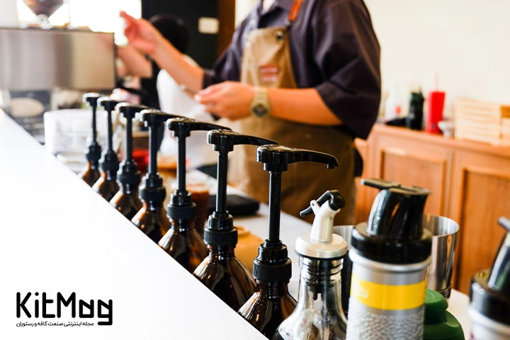 پمپ های غلیظ پاش مخصوص سیروپ که داخل یک کافی شاپ قرار گرفته اند و باریستا در حال درست کردن نوشیدنی های گرم و سرد است