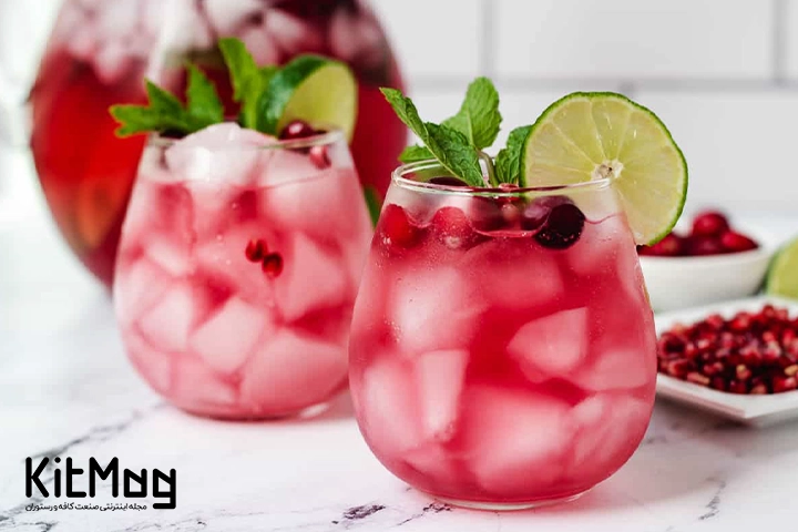 موکتل میموسای کرنبری (Cranberry Mimosa Mocktail) که داخل لیوان گرد پایه کوتاه ریخته شده است و با نعنا و لیمو تزئین شده است و در کنار آن انار ریخته شده است