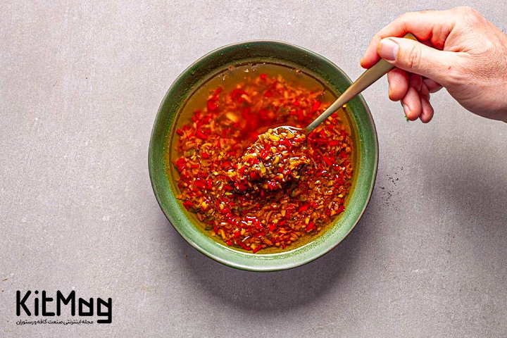 سس چیلی که داخل یک کاسه ریخته شده است و روی آن با روغن زیتون پر شده است و یک تستر در حال برداشتن آن با یک قاشق می باشد